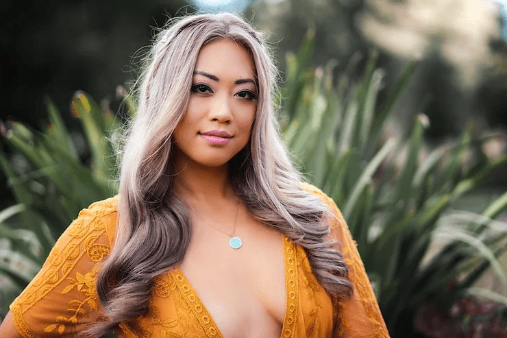 A young woman with long blonde hair wearing a yellow lace dress and a subtle necklace, posing in an outdoor setting with blurred greenery in the background.