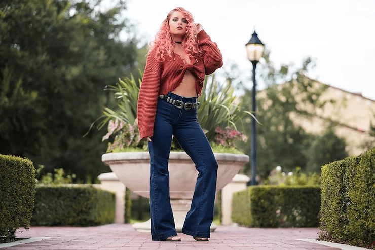 A woman with pink hair stands confidently in a garden, wearing blue jeans and a cropped orange sweater. a lamppost and trimmed hedges are in the background.