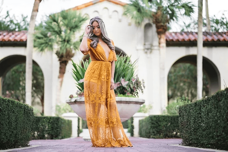 A woman in a flowing yellow lace dress stands confidently on a pathway, surrounded by lush greenery and elegant arches, with one hand adjusting her long hair.