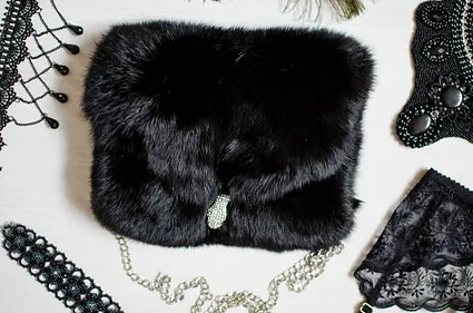 A black fur clutch with a jeweled clasp surrounded by various elegant accessories including lace, beads, and feathers on a white background.