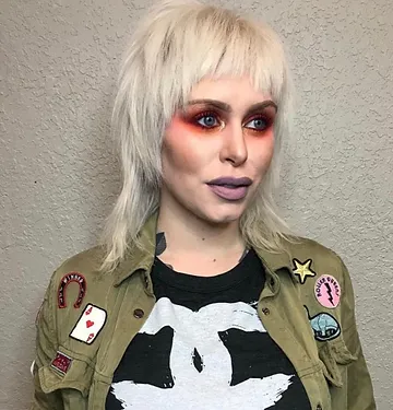 A woman with platinum blonde hair styled in a shag cut, wearing dramatic red eyeshadow and purple lipstick. she is dressed in a camouflage jacket with various patches and a graphic t-shirt.