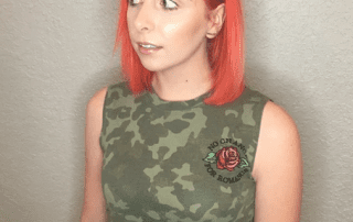 A woman with bright orange hair and a camo tank top, featuring a rose and text design, looks away from the camera, standing against a textured, neutral-colored wall.