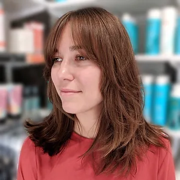 A woman with medium-length brown hair and bangs, wearing a red top, smiles gently while standing inside a store with blue bottles in the background.