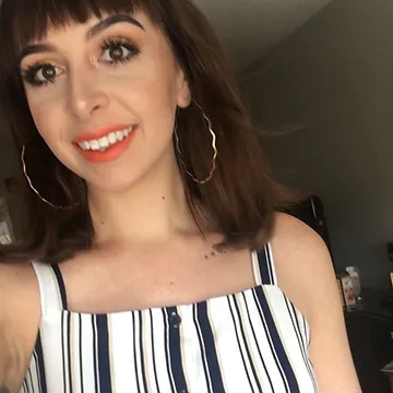 A woman with shoulder-length brown hair and bangs, wearing orange lipstick and large hoop earrings, smiles in a room. she has on a striped, sleeveless top.