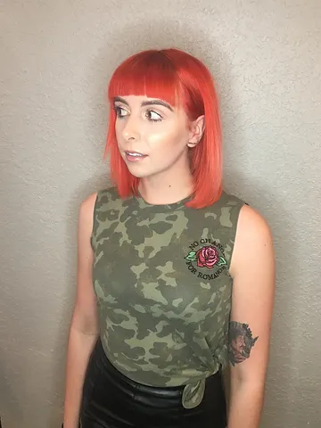 A woman with bright orange hair in a bob cut looks to the side, wearing a camo tank top with a rose emblem, paired with a black skirt. she has a visible tattoo on her right arm.