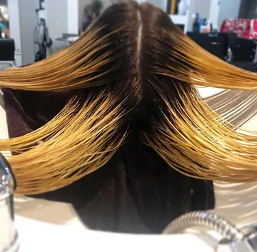 A woman at a hair salon with her hair splayed across the back of a salon chair, showcasing a multi-toned coloring effect, with shades ranging from dark at the roots to light blonde at the tips.