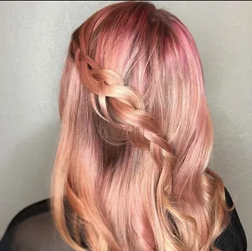 A woman with pink-toned hair styled into an intricate side braid, showing a gradient from deep rose at the roots to a lighter blush at the tips.