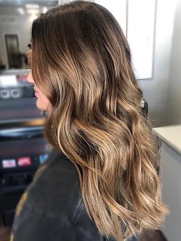 A woman in a salon chair, viewed from behind, showing her long, wavy hair with a glossy balayage blend of brunette and blonde highlights.