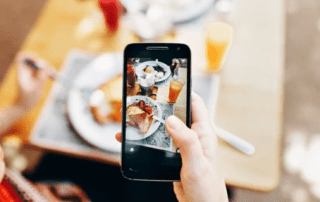 A person's hand holding a smartphone taking a photo of a breakfast spread with orange juice on a wooden table.