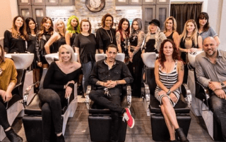 Group of people, including men and women of various ages and styles, posing in a modern salon with salon chairs and mirrors in the background. some are seated and some are standing.