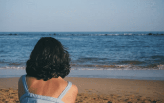 A woman with short, black hair, seen from behind, gazes at the ocean while sitting on a sandy beach. she is wearing a light blue dress.