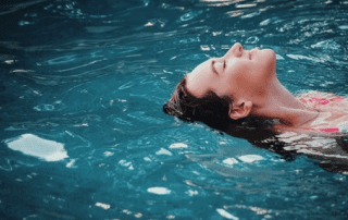 A woman peacefully floating on her back in dark blue water, her face partially submerged, with her eyes closed in relaxation.