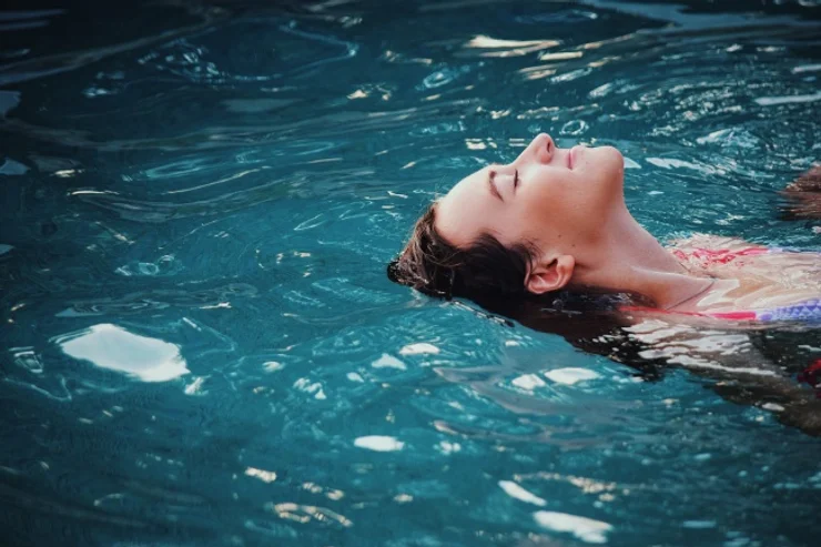 A woman relaxes by floating on her back in a serene blue pool, her eyes closed and face partially submerged in water, conveying a sense of peacefulness.