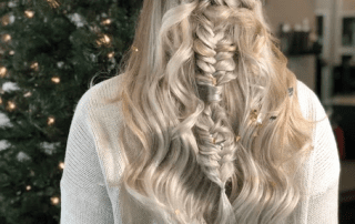 Blonde woman with a detailed braided hairstyle and a floral crown, viewed from behind, with a decorated christmas tree and twinkling lights in the background.