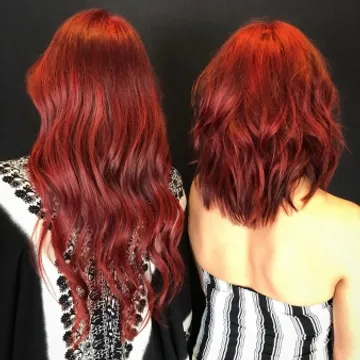 Two women with vibrant red hair, one with long wavy hair and the other with a short tousled hairstyle, standing back-to-back against a black background.