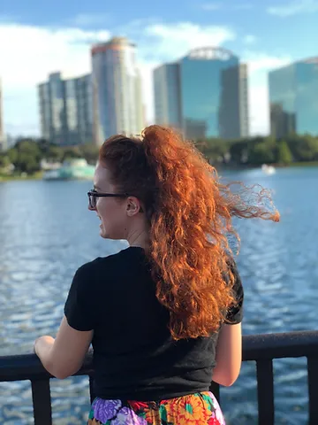 A woman with curly red hair wearing sunglasses and a black shirt standing by a railing, looking at a city skyline across a body of water.