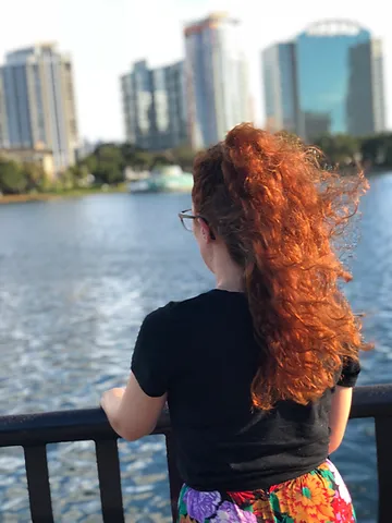 A woman with curly red hair gazes at a city skyline across a river from a bridge railing, wearing a colorful skirt and black top.