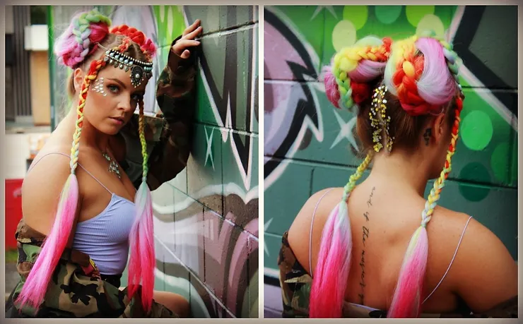 A woman with vibrant rainbow-colored hair styled in braids and buns, adorned with beads and flowers, poses confidently against a graffiti-covered wall.