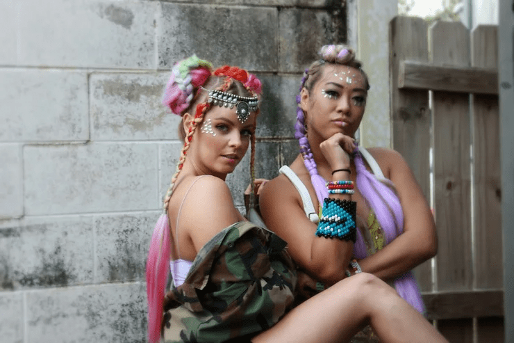 Two women with colorful, elaborately styled hair and bold makeup sit back-to-back. they wear vibrant accessories and a mix of modern and traditional attire, posing confidently near a stone wall.