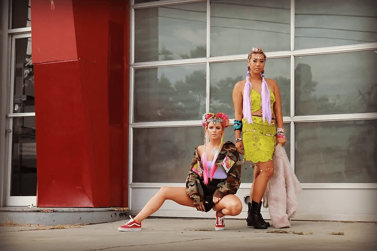 Two women in vibrant, eclectic outfits pose confidently in front of a modern building with red columns and large windows. one stands and one squats, both wearing colorful headpieces and accessories.