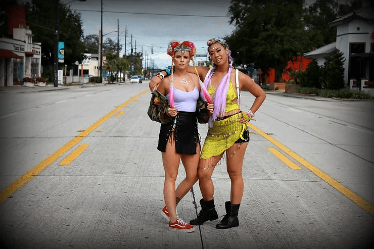 Two women standing confidently in the middle of an empty street, wearing bold, colorful outfits and accessories, with buildings lining the road under a cloudy sky.