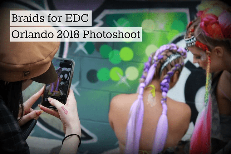 A person captures a photo of two women with colorful braided hairstyles, featuring the theme 'braids for edc orlando 2018 photoshoot’, against a blurred background of green lights.