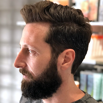 Side profile of a man with a styled, voluminous quiff and a full, well-groomed beard, set against a blurred indoor background.