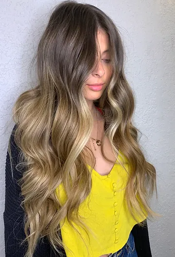 A woman with long, wavy hair featuring a smooth transition from dark roots to light blonde tips, partially covering her face, wearing a yellow top.