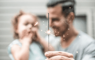 A man and a young girl enjoy a sparkling firework together, blurred house in the background. the child appears delighted by the bright sparks.