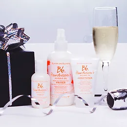 A festive display featuring haircare products with a glass of champagne, a wrapped gift box, and ribbon on a white surface.