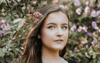 A young woman with light brown hair gazes upward among purple flowers and green foliage in a tranquil garden setting.