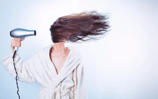 A person in a white robe using a hairdryer, causing their long, brown hair to dramatically blow to the side against a light blue background.