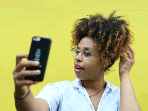 A woman with curly hair taking a selfie with her smartphone against a yellow background. she is wearing glasses and a white t-shirt.