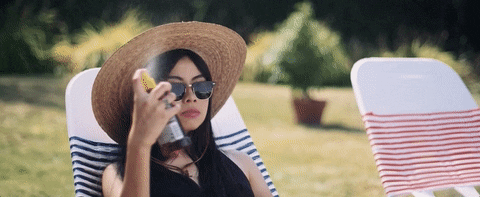 A woman wearing a wide-brimmed hat and sunglasses lounges on a sun chair in a garden, intently looking at her mobile phone.