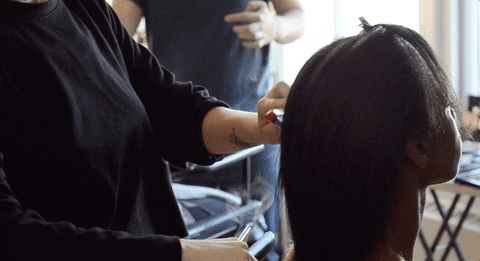 A hairstylist secures a section of a woman's hair with a bright red hair clip, preparing to style it in a busy salon setting.