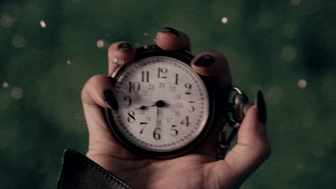 A close-up of a hand holding an open pocket watch, showing a classic white face with black roman numerals. snowflakes gently fall around in a dark, blurred background.