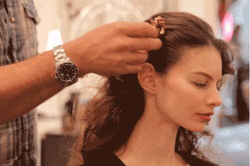 A woman in a salon receives a hairstyle from a stylist who is securing her hair into an updo using bobby pins.