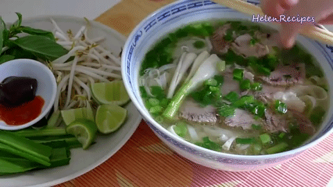 A bowl of traditional vietnamese pho with sliced beef, noodles, and herbs, served with a side of fresh basil, bean sprouts, lime, and hoisin sauce. a hand is adding ingredients to the soup.