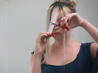A woman with her hair up is cutting her bangs in front of a mirror, using red scissors. her expression is focused.