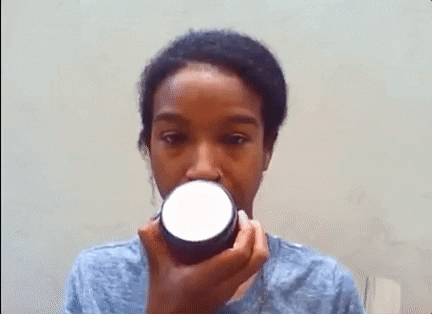 A woman examines the label of a jar she is holding up close to her face, against a plain background.