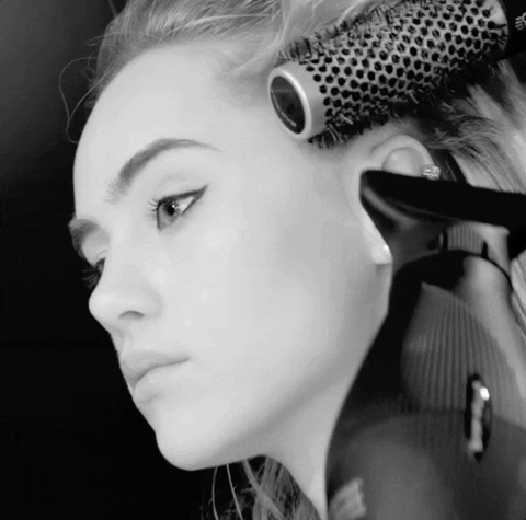 A black and white close-up of a woman getting her hair styled using a round brush and hairdryer, focusing on her profile as she looks away.