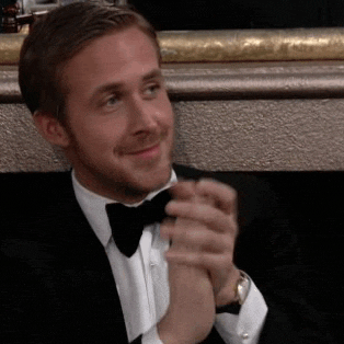 A man in a black tuxedo and bow tie, sitting and smiling while clapping his hands. he has blond hair and a neatly groomed beard.
