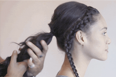 A woman with partially braided hair gazes to the side as a hairstylist continues to braid the rest of her hair.