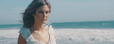 A woman in a white dress stands on the beach, with wind blowing her hair, looking away while the waves crash in the background.