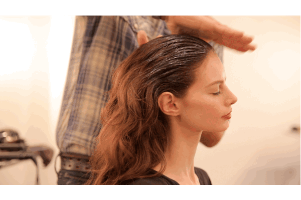 A hairstylist applies product to a woman's long hair at a salon, enhancing texture and shine. the woman, with eyes closed, appears relaxed during the styling session.