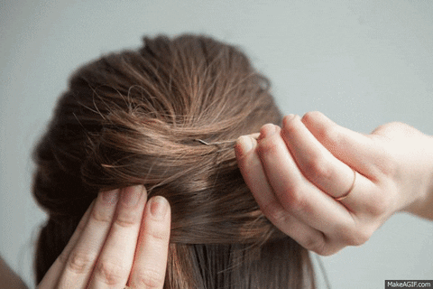 A woman's hands are styling a section of brown hair into a braided updo on the back of her head. the focus is on her hands and the hair being styled.