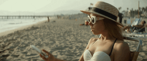 A woman in a sunhat and sunglasses sits on a beach chair, holding a smartphone with the ocean and a pier in the background.