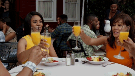 Two women toasting with mimosas at an outdoor brunch table filled with colorful dishes, surrounded by other diners in a lively atmosphere.