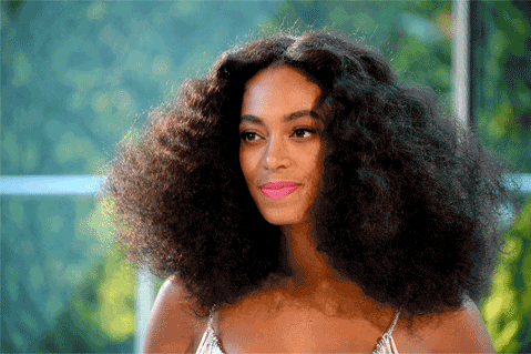 A woman with voluminous curly hair and a subtle smile is wearing a light-colored top, sitting outdoors with green foliage in the background.