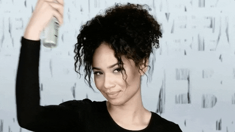 A woman with curly hair smiles at the camera while playfully spraying her hair with a product from a canister in her hand.